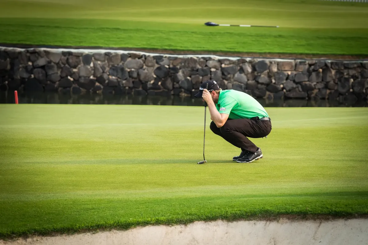 A golfer crouches on a lush green golf course, concentrating on putting strategy.